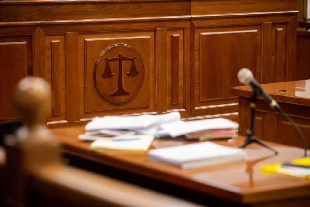 A wooden courtroom bench with the scales of justice carved on it, a table in front covered with documents, books, a notepad, and a microphone—reminding you of the importance of choosing the right criminal defense attorney.